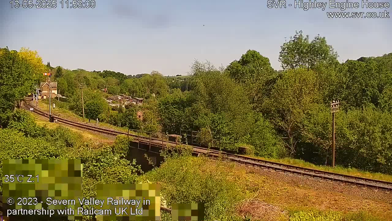 A sunny view of a railway track winding through a lush green landscape, with a small station visible in the distance.