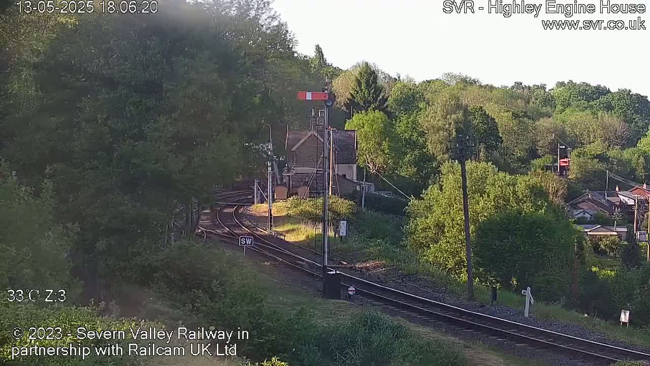 A sunny view of a railway line curving through a tree-lined hillside, with a small stone building and signal near the tracks.