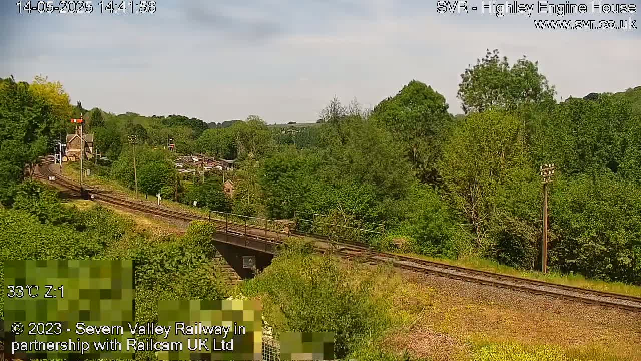 A railway line stretches through a verdant landscape on a sunny day, passing over a bridge near a small building.