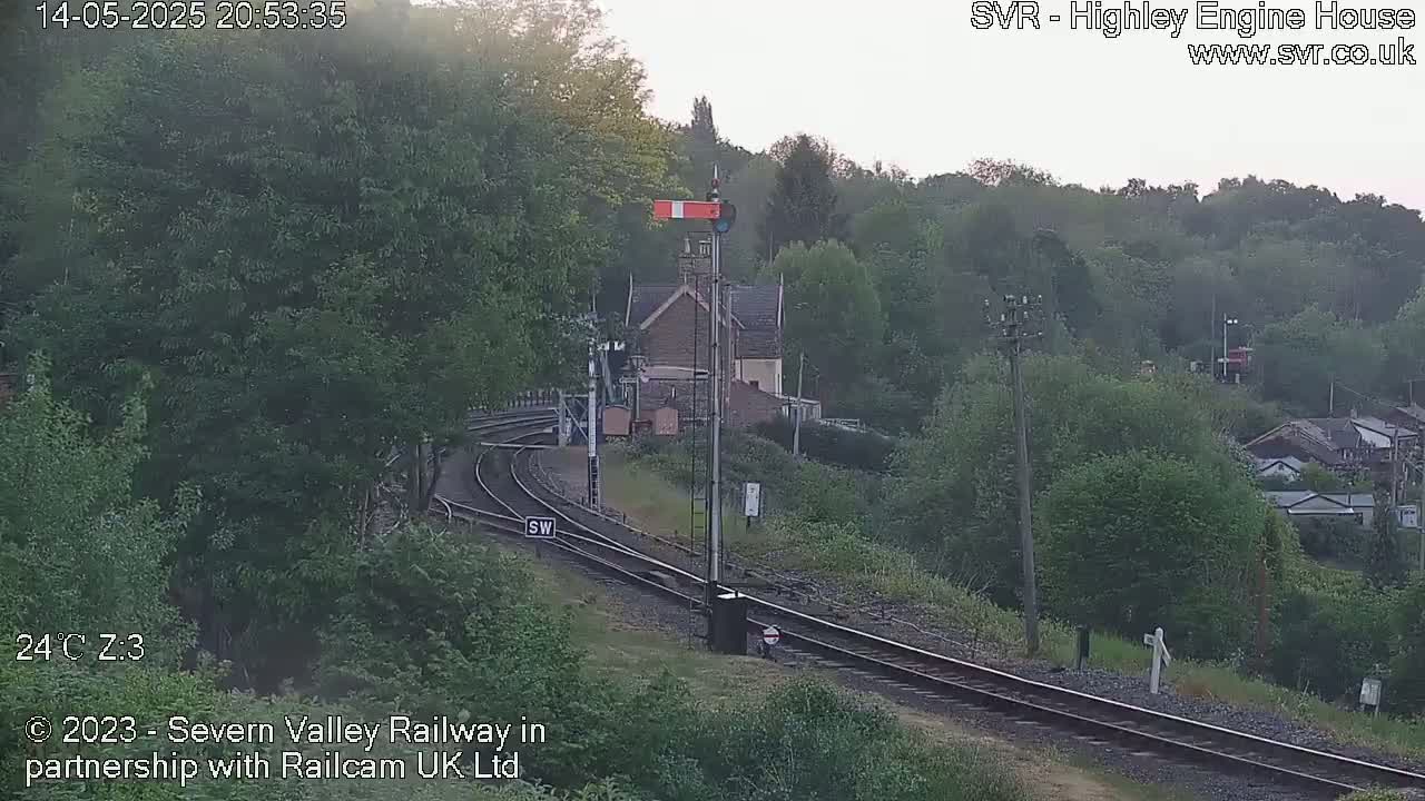 A railway track curves through a lush green landscape, under a hazy sky, near a small stone building.