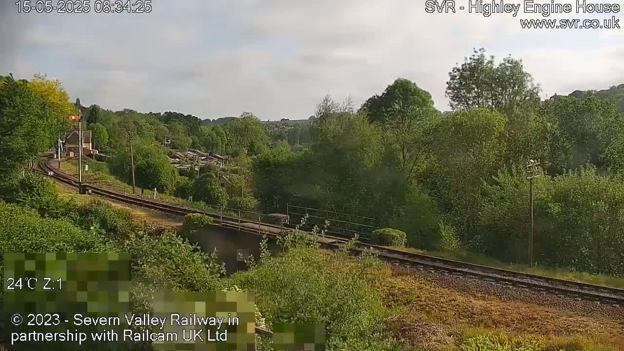 A train passes over a bridge on a railway track through a lush green landscape under a partly cloudy sky.