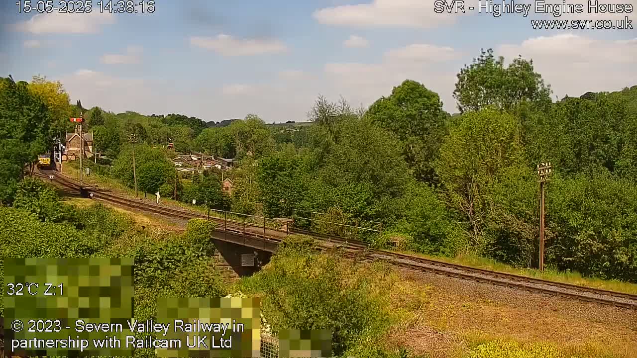A train approaches a railway bridge under a partly cloudy sky on a sunny day, passing through a lush green landscape.