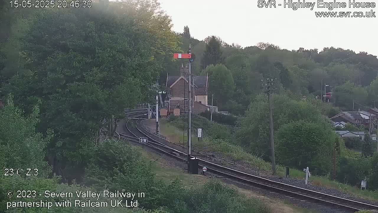 A railway line curves through a green, wooded area near a stone building under an overcast sky.