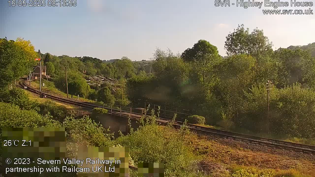 A sunny view of a railway track winding through a lush green landscape with trees and buildings in the distance.