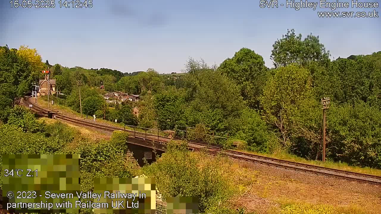 A sunny day reveals a railway track curving through a lush green valley, passing over a bridge near a small village.