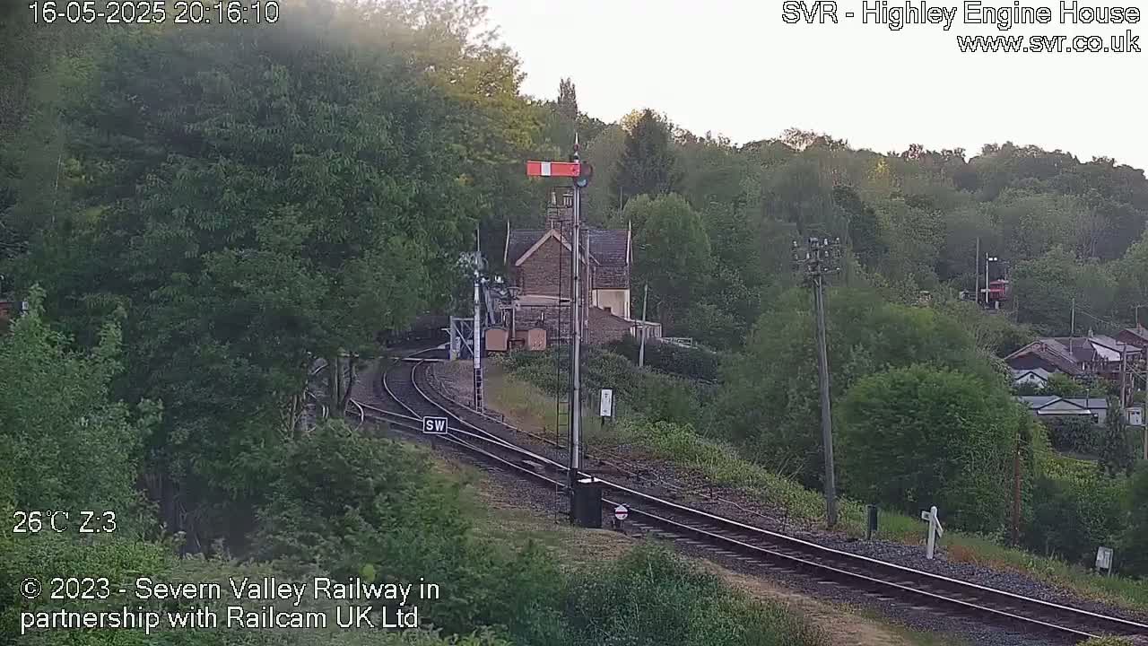 A train track curves through a lush green landscape at dusk, passing a stone building near a signal and alongside a hill dotted with houses.