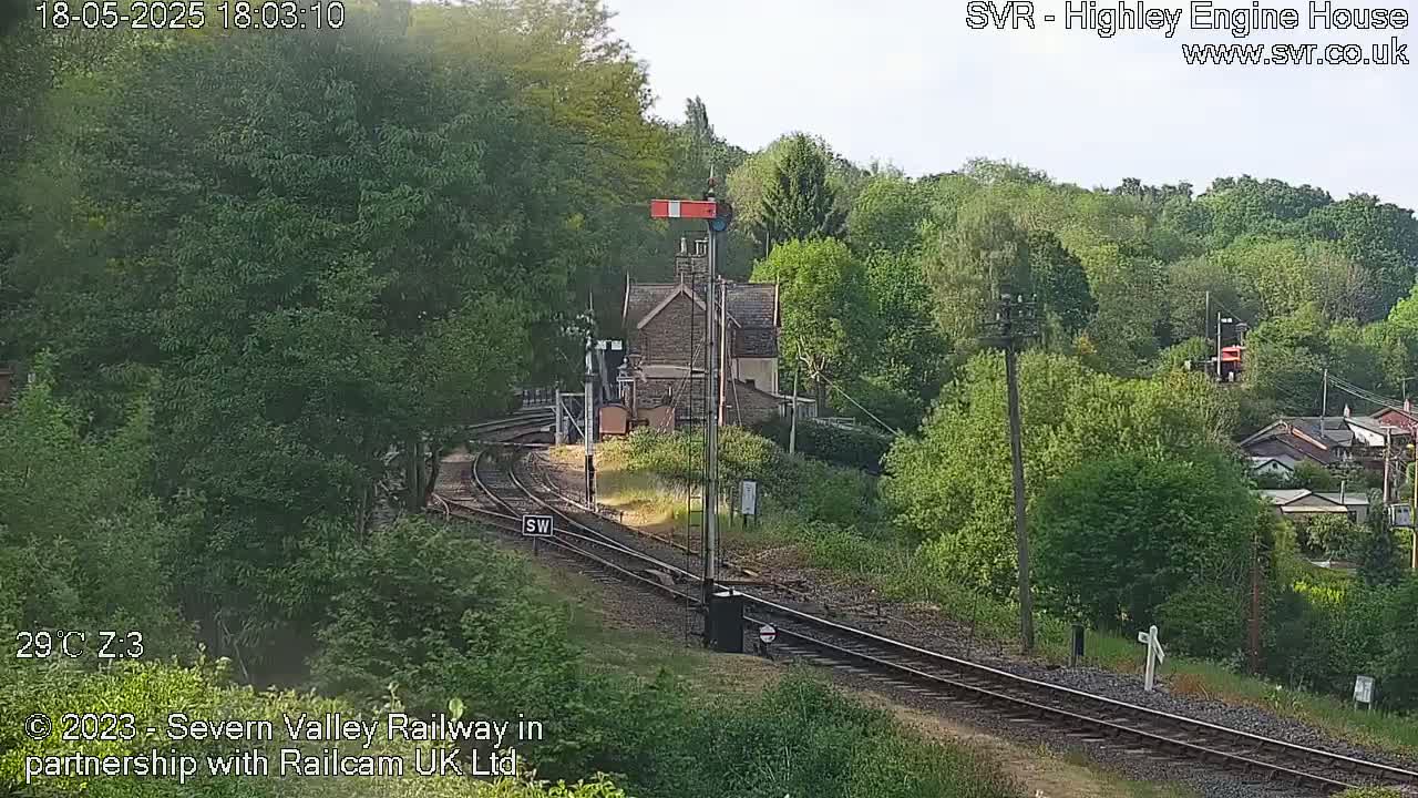 A railway line curves through a green, leafy landscape on a sunny day, passing a small stone building near a signal.