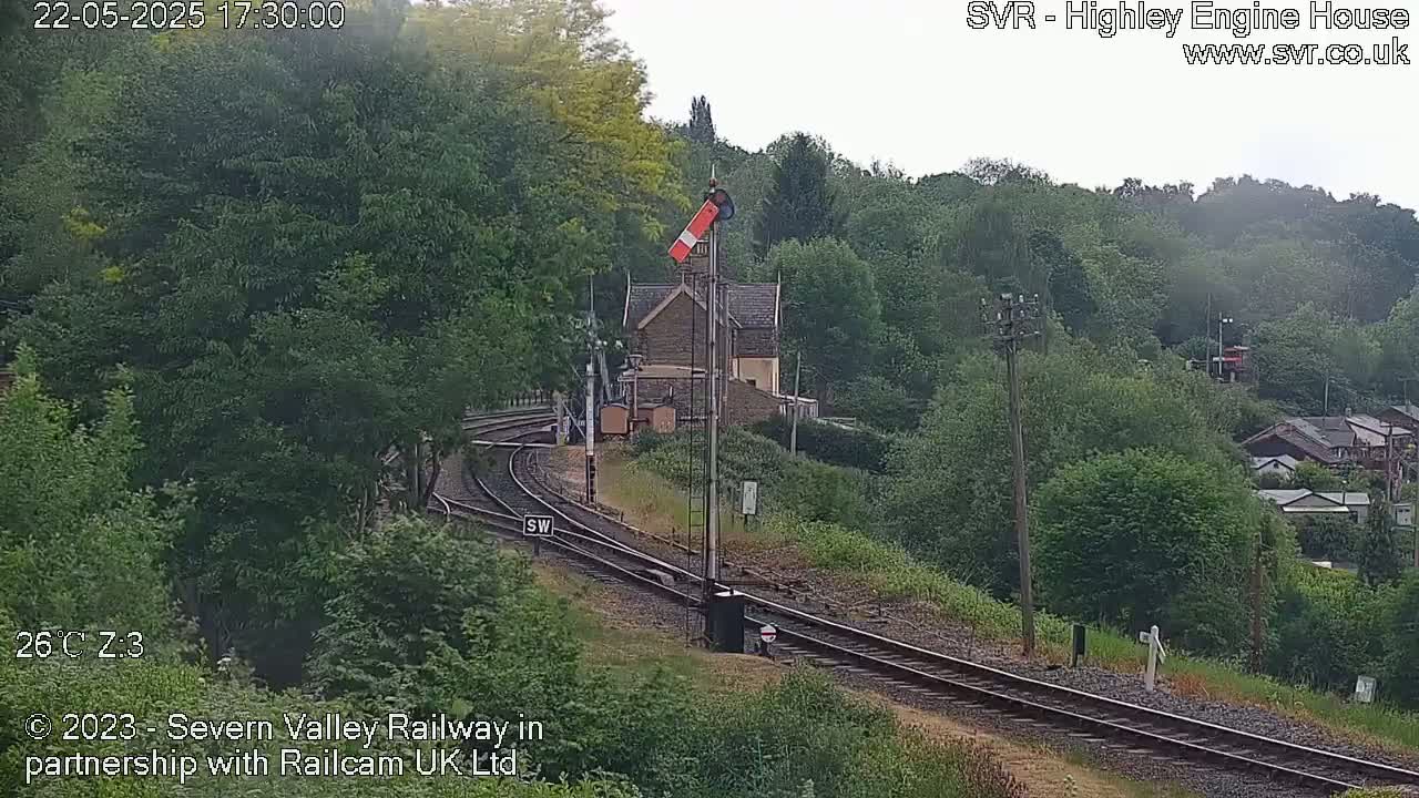 A railway line curves through a lush green landscape, passing a stone building near a signal, under an overcast sky.