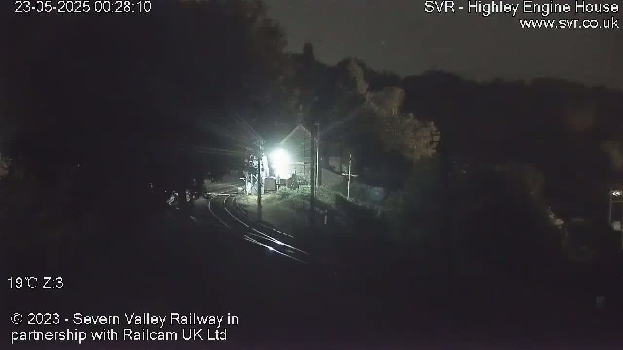A dimly lit nighttime scene shows a small building next to train tracks, surrounded by dark trees under a clear sky.