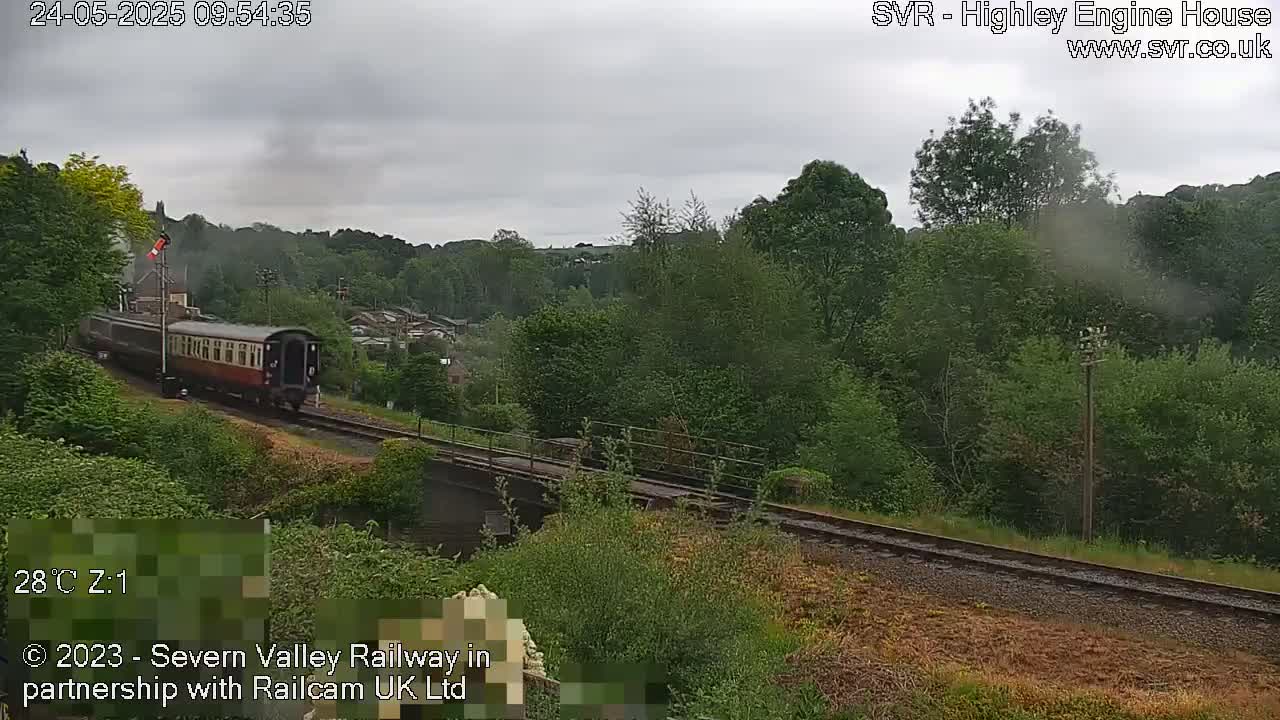 A passenger train is traveling across a bridge through lush green woodland under an overcast sky.