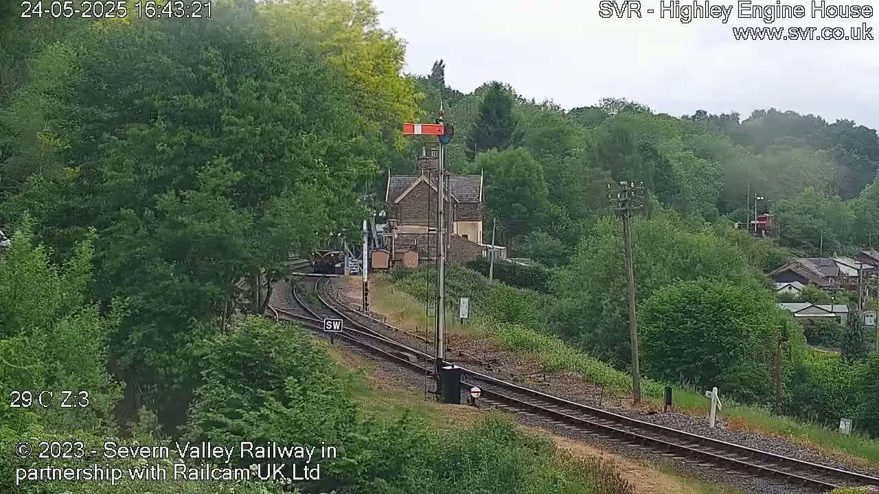A train is approaching a station nestled in a lush green hillside under a partly cloudy sky.