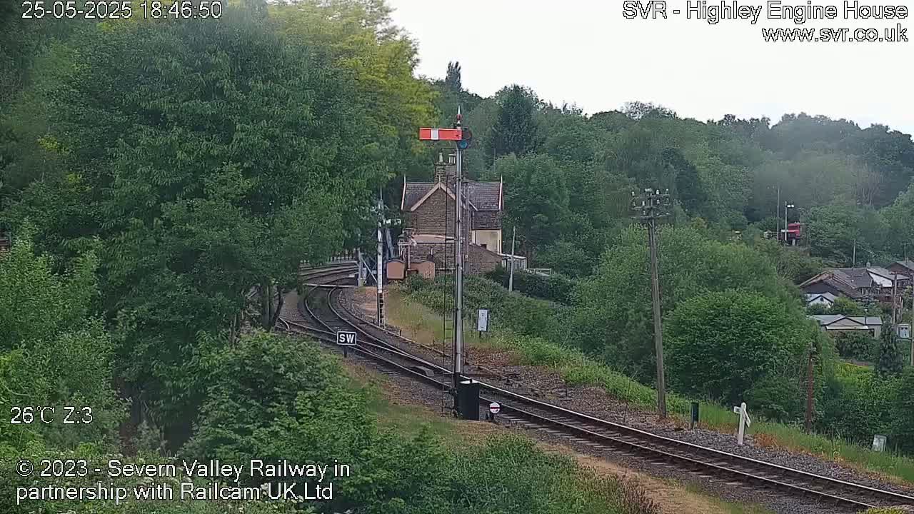A railway track curves through a lush green landscape, passing a small stone building under a partly cloudy sky.