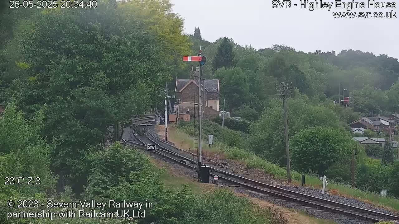 A train track curves through a lush green landscape on an overcast day, with a small stone building and signal visible near the tracks.