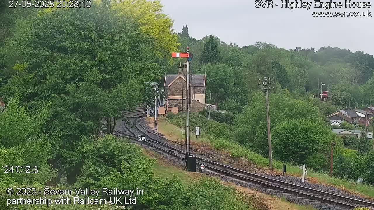 A railway line curves through a lush green valley, passing a small stone building under an overcast sky.