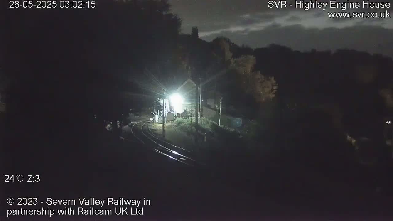 A nighttime overhead view of a dimly lit train station with train tracks, surrounded by dark trees under a cloudy sky.
