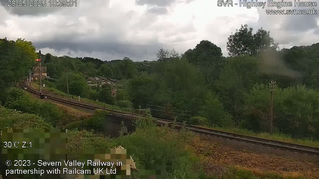 A railway track curves through a lush green landscape under a cloudy sky.