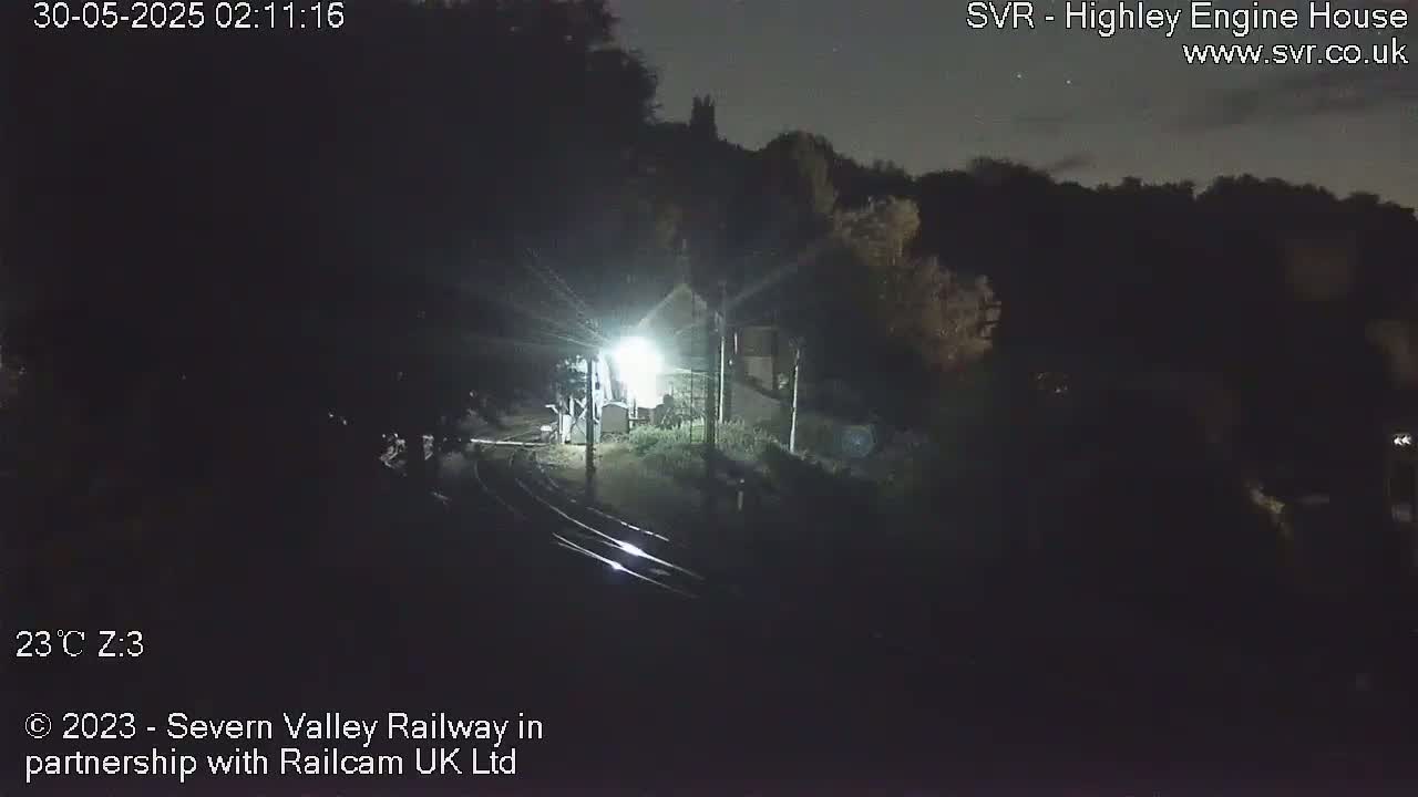 A nighttime, dimly lit view of train tracks curving toward a building, surrounded by dark trees under a clear sky.