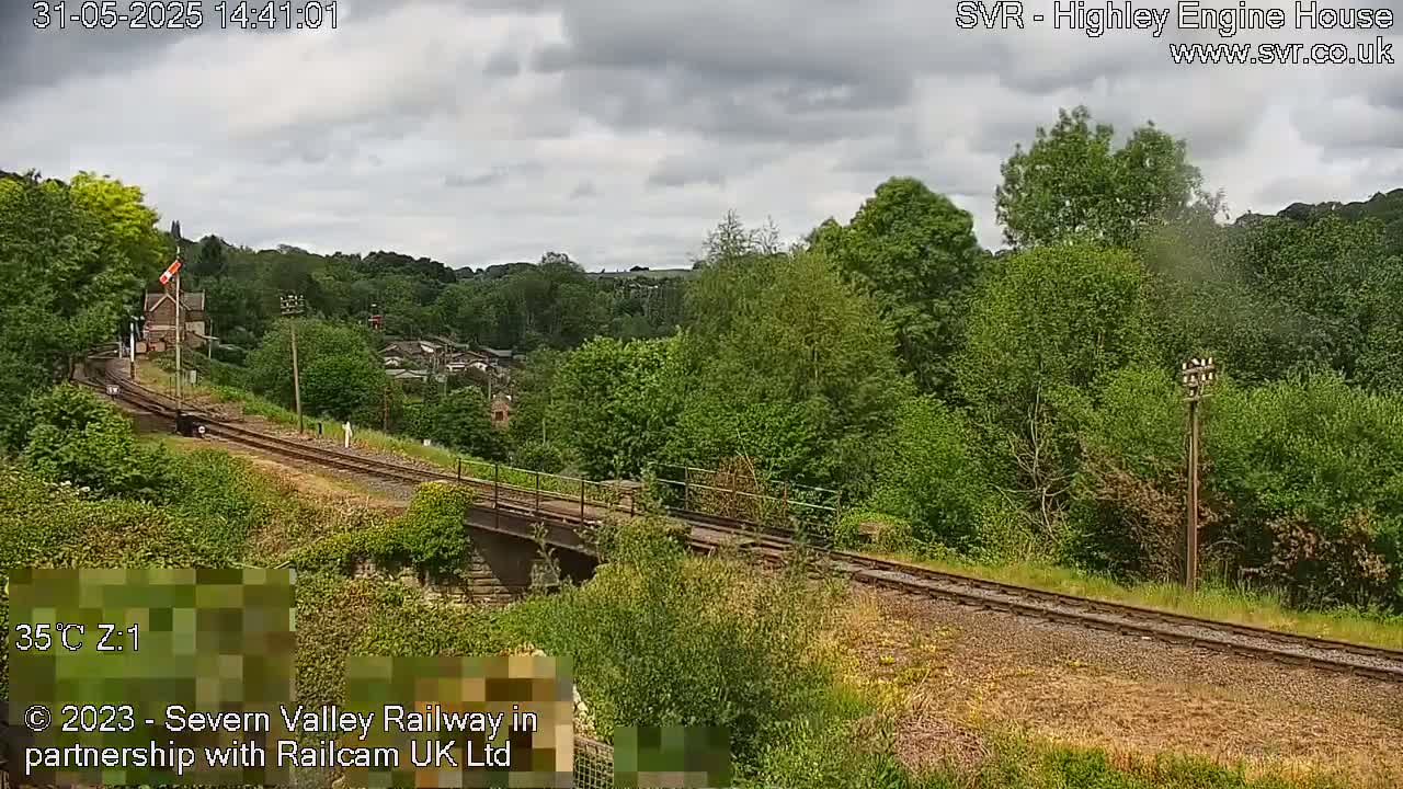 A train travels along railway tracks that cross a bridge, surrounded by lush green trees and vegetation under a partly cloudy sky.