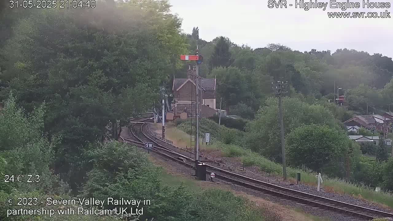 A view of a railway line curving through a green, wooded landscape under an overcast sky.