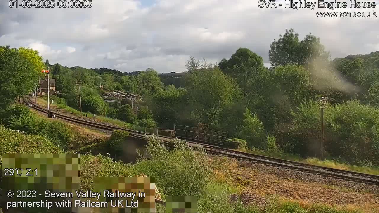 A train passes along railway tracks that wind through a lush green valley under a partly cloudy sky.