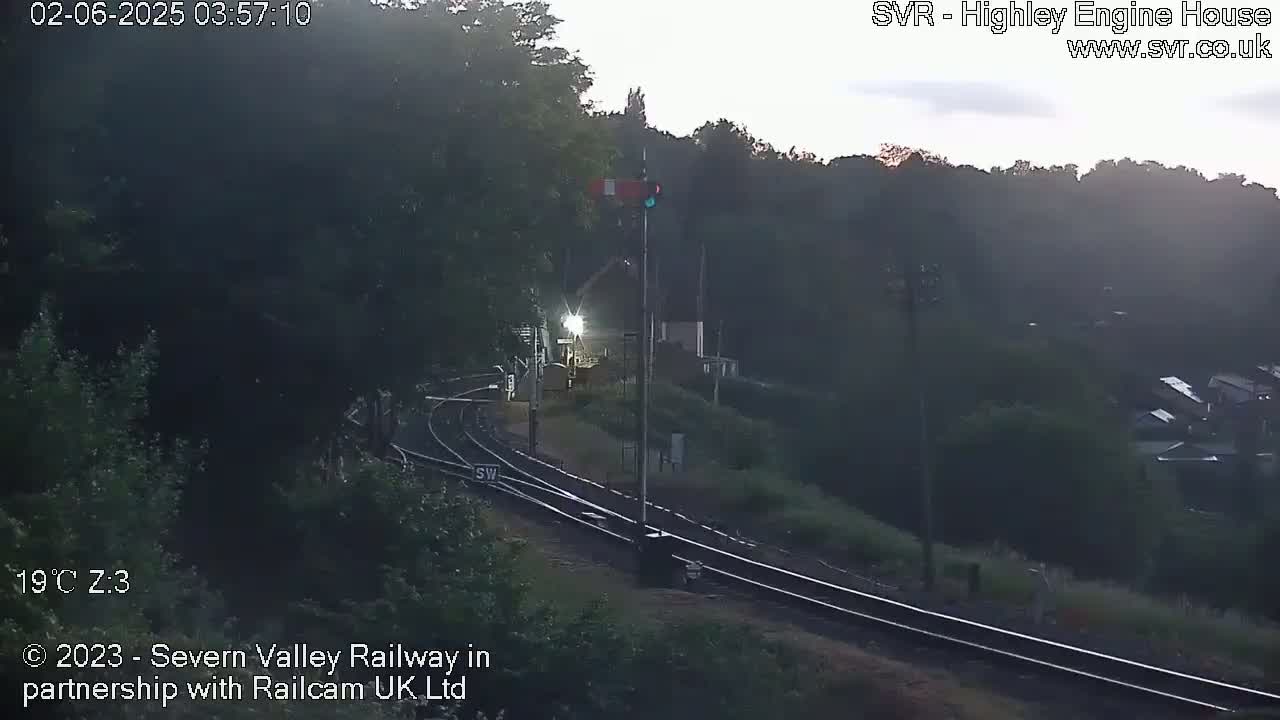 A dimly lit train station with railroad tracks and a signal light at dawn, surrounded by trees and greenery.
