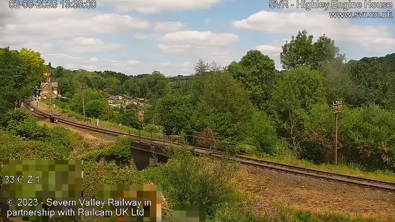 A partly cloudy day reveals a railway track curving through a lush green landscape, passing over a bridge near a small building.