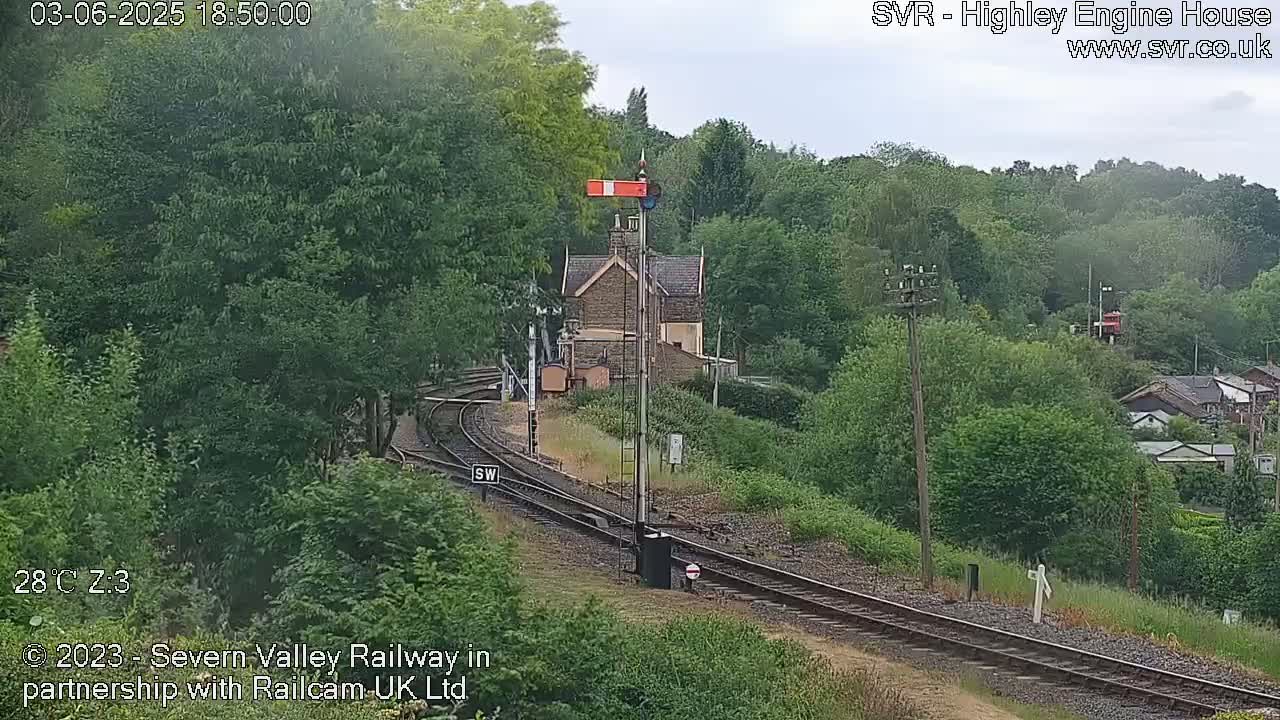 A railway line curves through a green, wooded area on an overcast day, with a small stone building visible near the tracks.