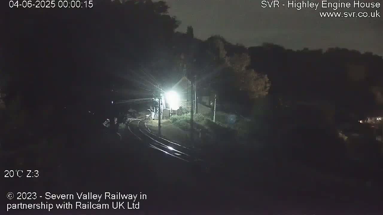 A nighttime, overhead view of a dimly lit train station with train tracks curving away from the camera, surrounded by dark trees and buildings under a clear sky.