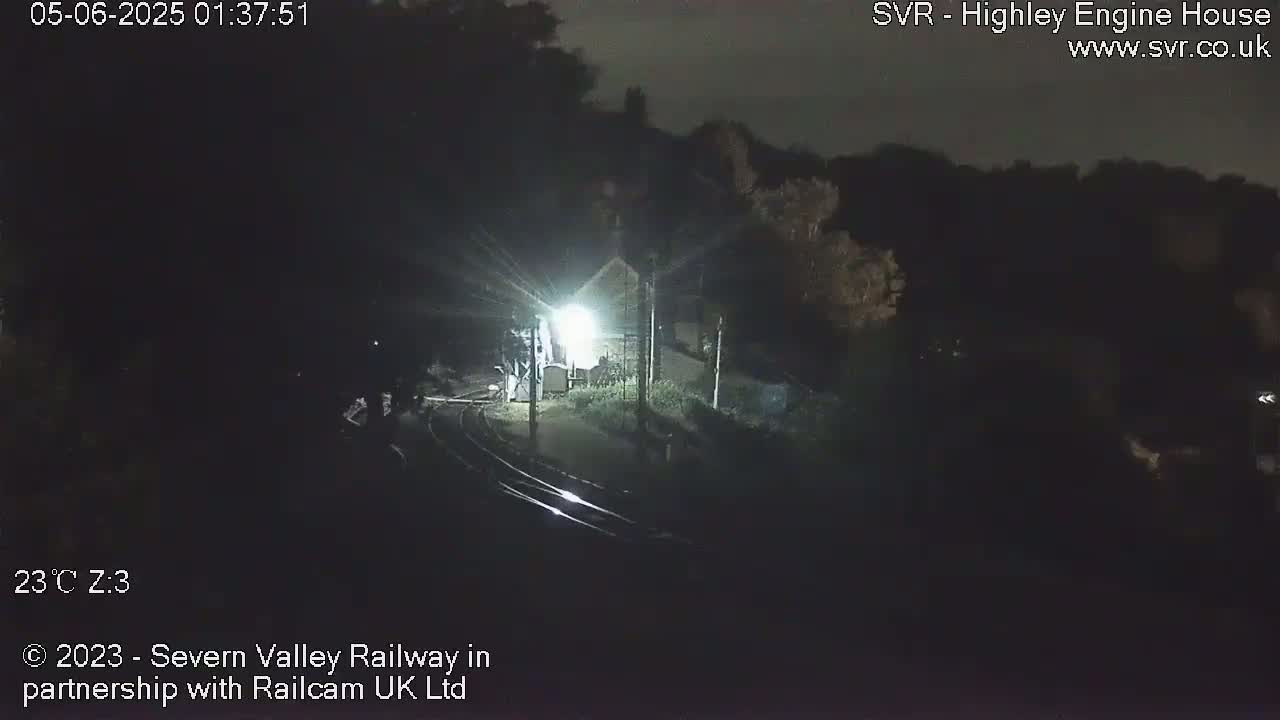 A nighttime, dimly lit view of train tracks leading to a building, surrounded by dark trees under a clear sky.