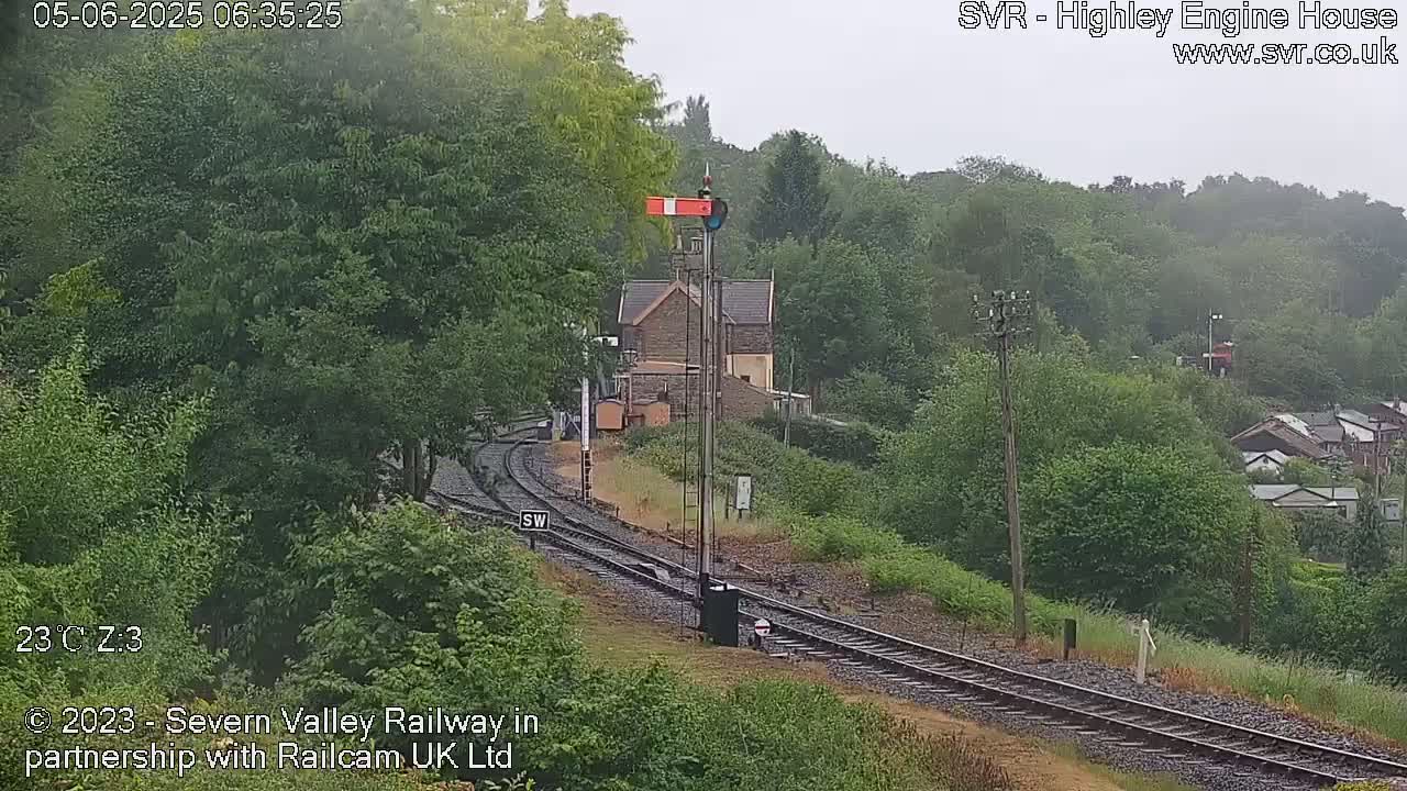 A railway track winds through a lush green landscape under an overcast sky, passing a small stone building.