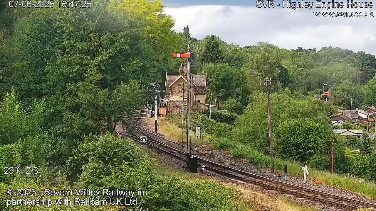 A railway track curves through a lush green landscape under partly cloudy skies, past a stone railway station building.