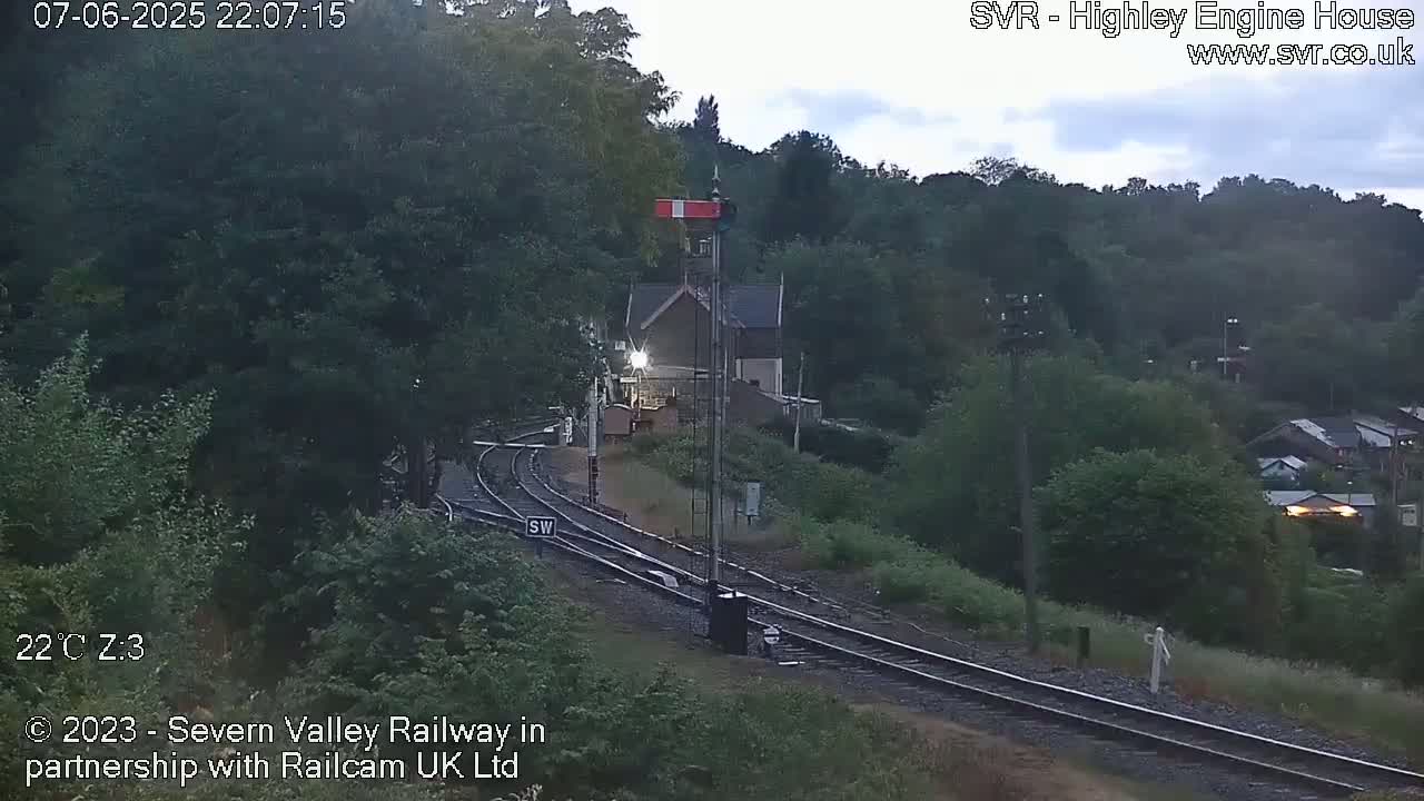 A nighttime view of a train station with two sets of tracks, surrounded by lush green trees under a cloudy sky.