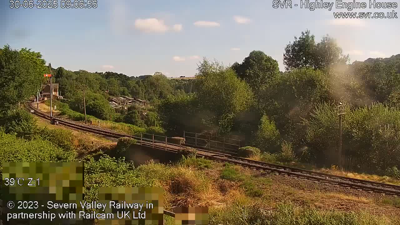 A train travels along a railway track that curves through a lush green valley under a partly sunny sky.