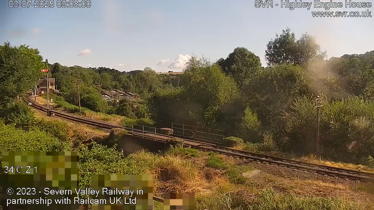 A sunny view of a railway track curving through a lush green landscape, passing over a bridge.