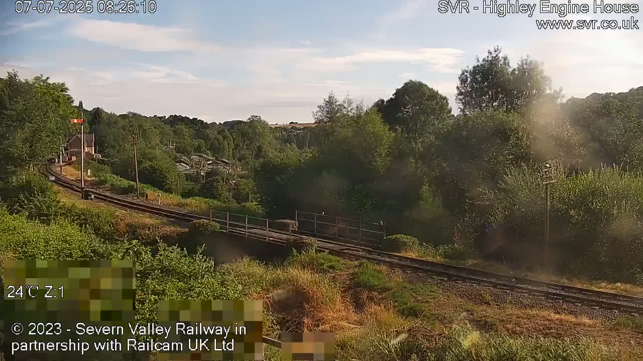 A train moves along a railway track that runs through a lush green landscape under a partly cloudy sky.