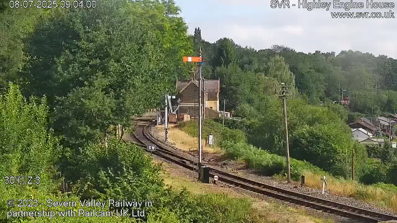 A train station building is visible beside a curving railway track that runs through a lush green landscape on a sunny day.