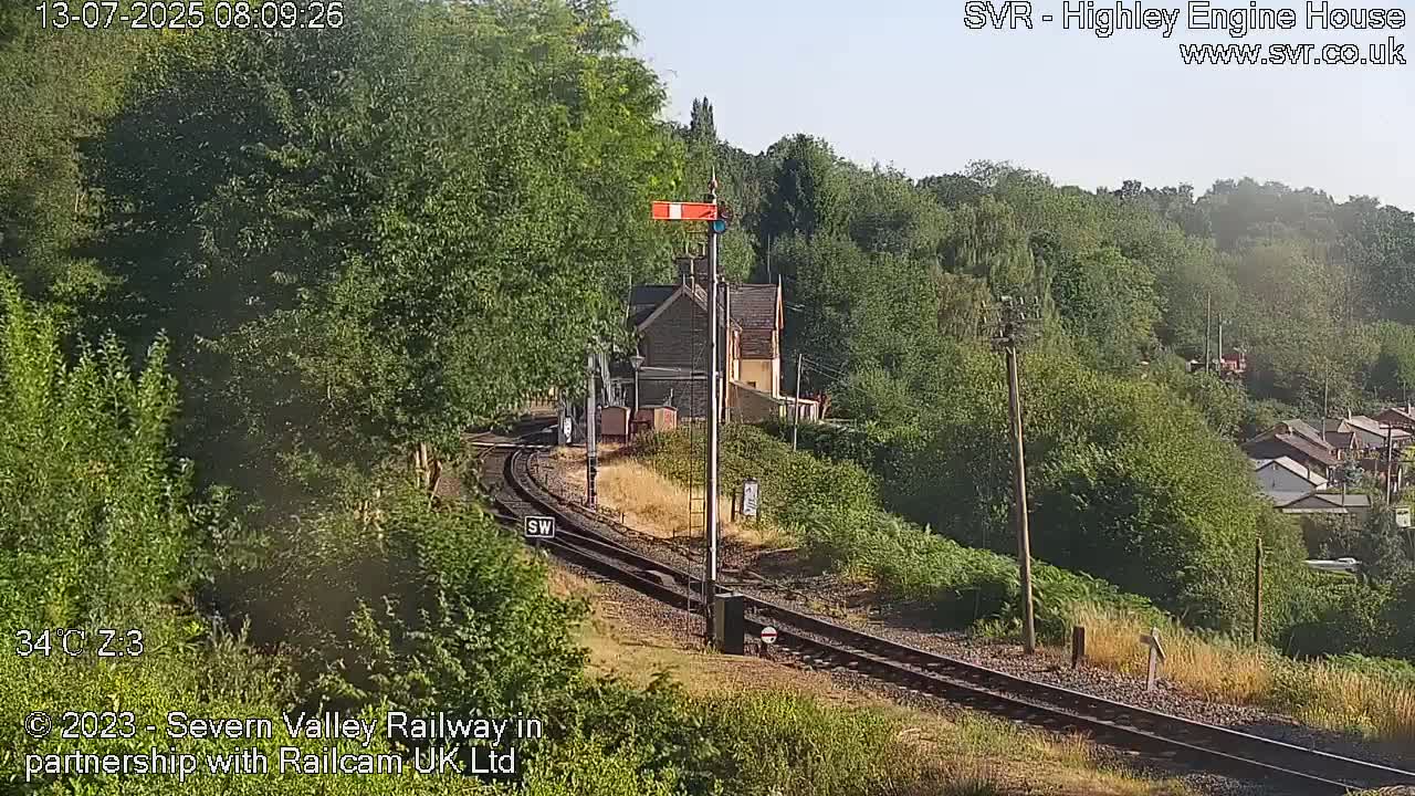 A sunny day reveals a railway track curving through lush green vegetation towards a small building, with houses visible on a hillside in the background.
