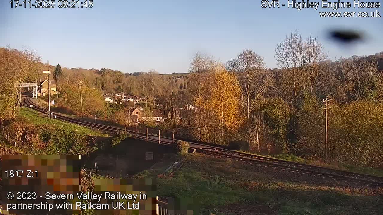 A sunny day reveals a scenic view of a railway line winding through a valley past a small station building and scattered houses, surrounded by trees with hints of autumn foliage under a clear blue sky.
