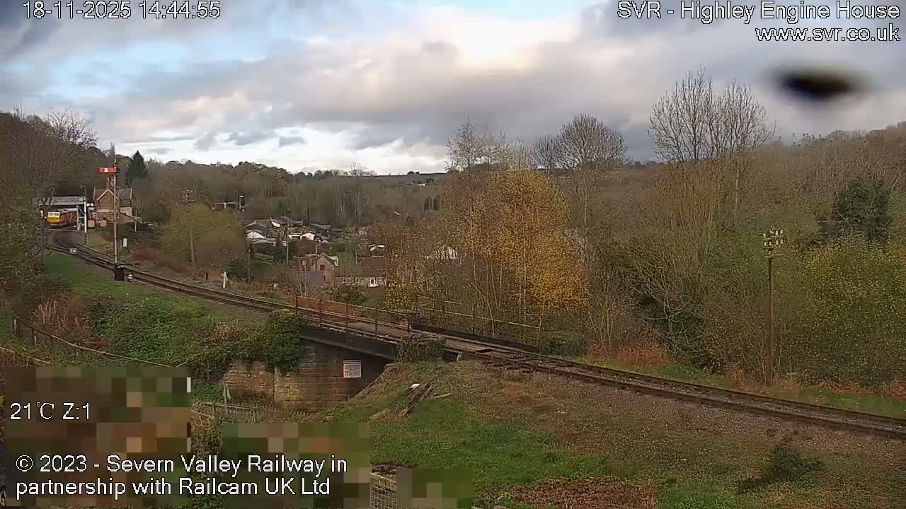 A sunny day reveals a scenic view of a railway line winding through a valley past a small station building and scattered houses, surrounded by trees with hints of autumn foliage under a clear blue sky.