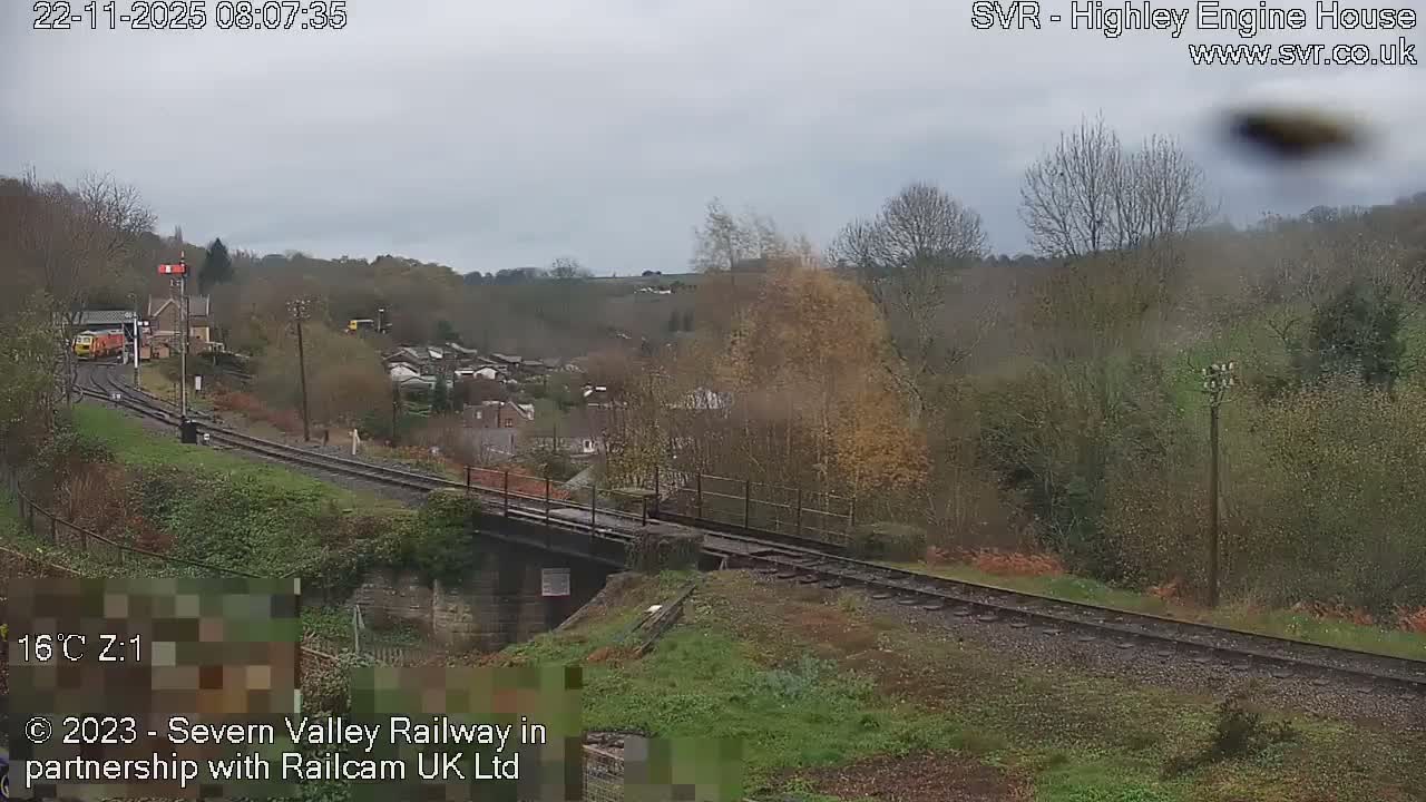 A yellow and orange train is visible on railway tracks curving towards a station and village nestled within a hilly landscape filled with trees, all under a dull, overcast sky.