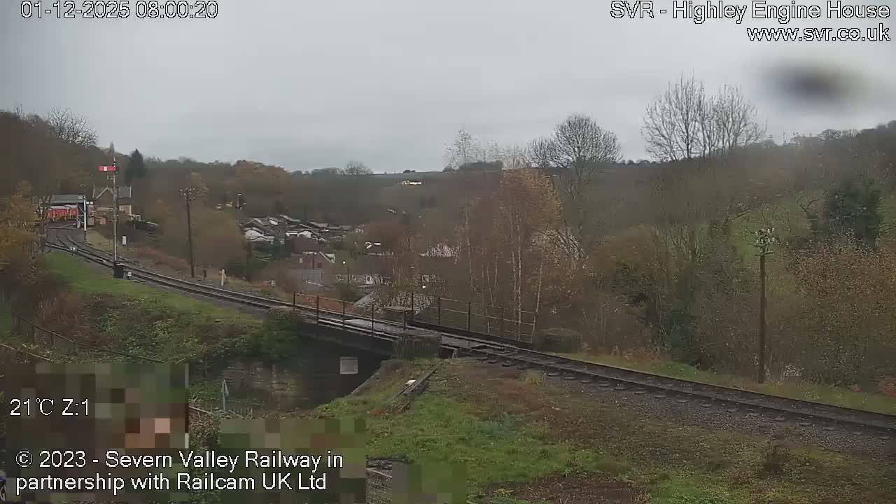 An overcast morning view shows a railway line with a red signal and a station building, curving over a bridge and through a hilly, tree-covered landscape dotted with houses.
