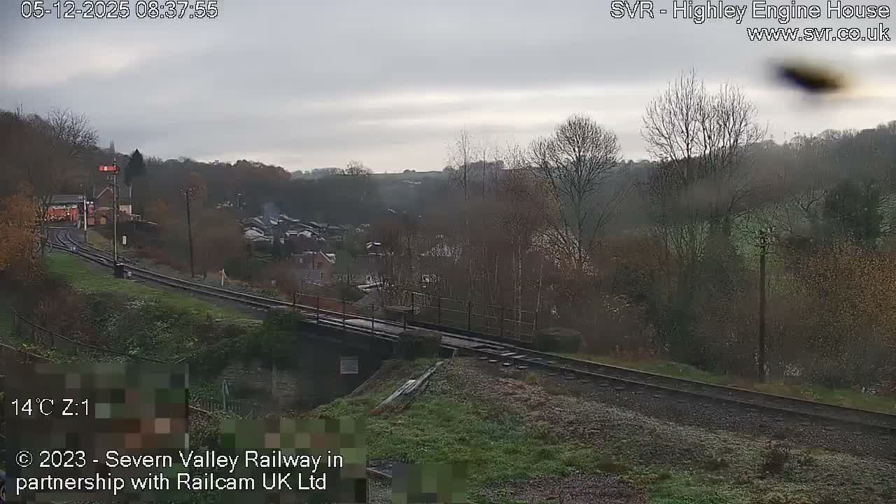 An overcast sky hangs over a valley landscape featuring curving railway tracks that cross a bridge, flanked by bare trees and distant buildings.