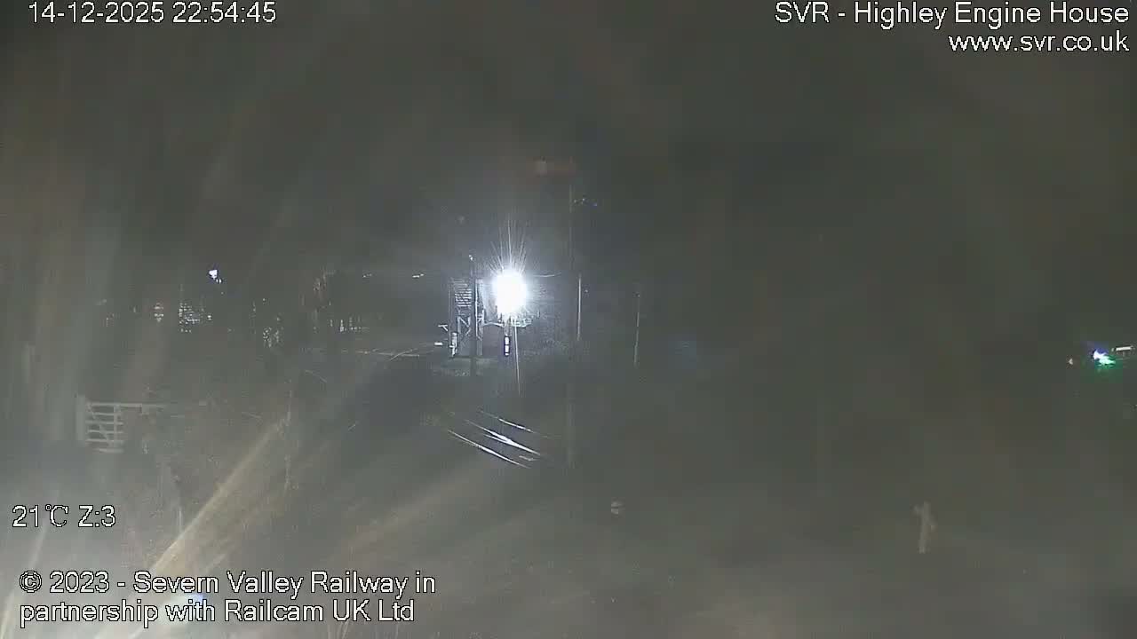 An overcast sky hangs over a valley landscape featuring curving railway tracks that cross a bridge, flanked by bare trees and distant buildings.