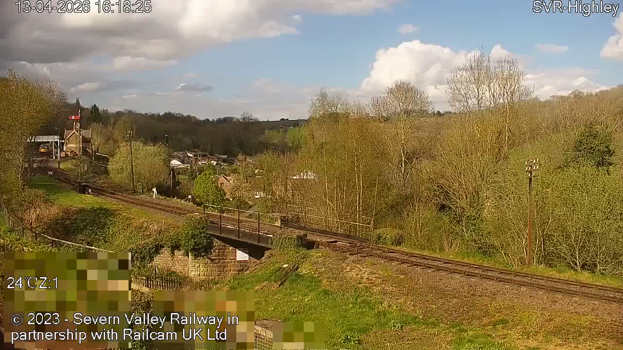 An overcast sky hangs over a valley landscape featuring curving railway tracks that cross a bridge, flanked by bare trees and distant buildings.