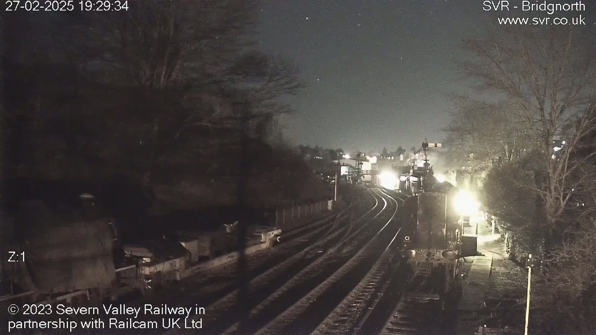 A nighttime view of train tracks curving through a dark, sparsely lit area with bare trees under a clear, starlit sky.