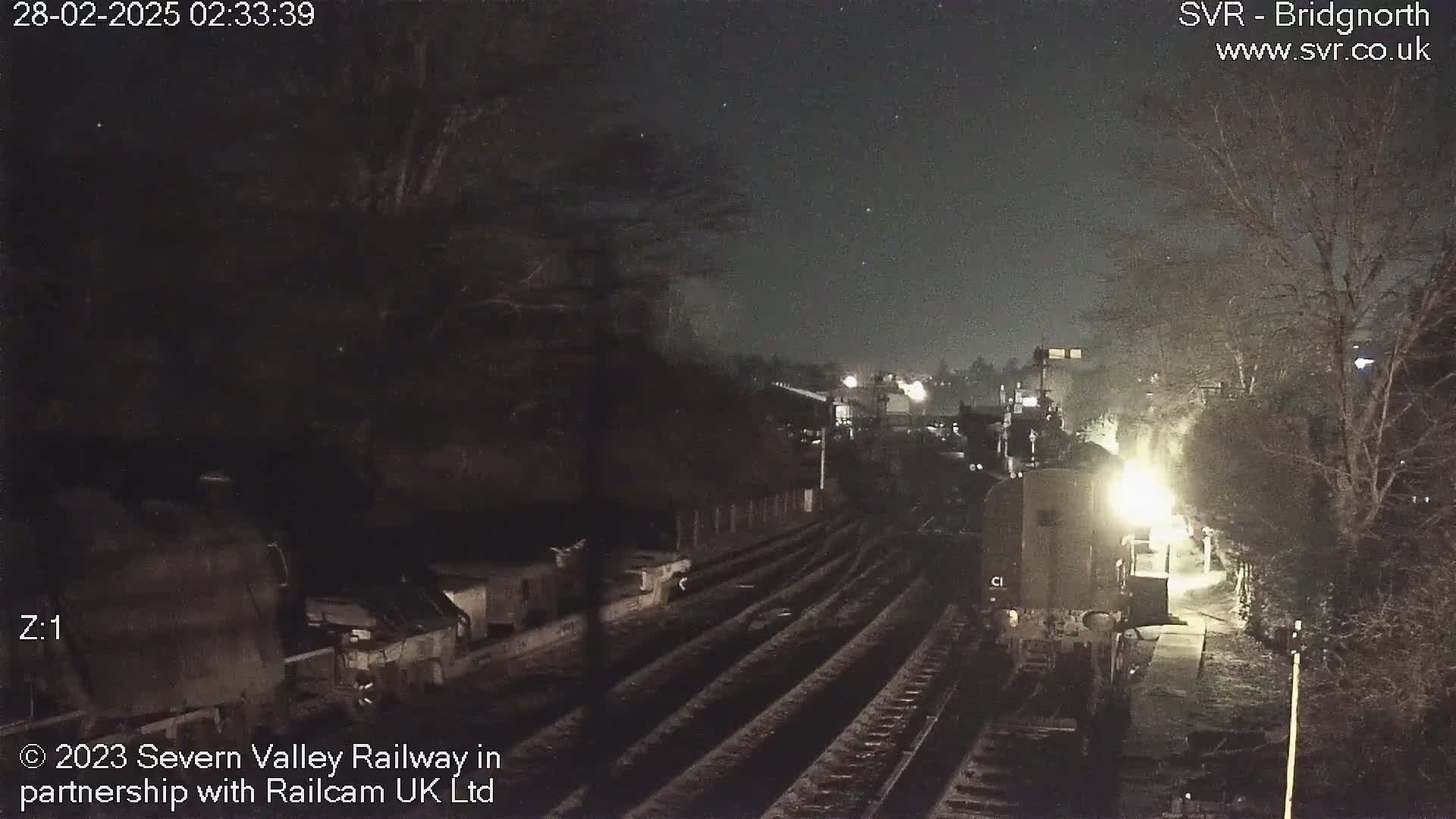 A nighttime view of a train at a station, illuminated by its headlights, under a clear, starry sky.