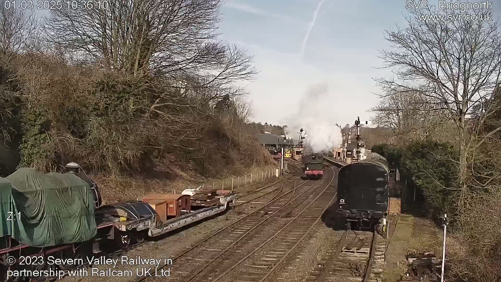 Two steam trains are traveling on a railway track on a sunny day, passing a freight train carrying wooden crates.