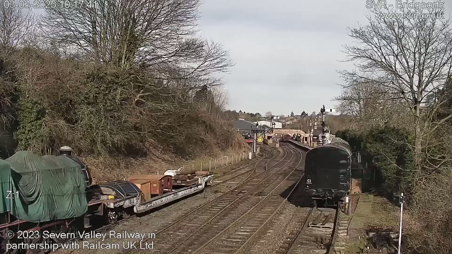 Two trains are on a railway track on a partly sunny day, one covered and carrying cargo, the other a dark-colored locomotive.