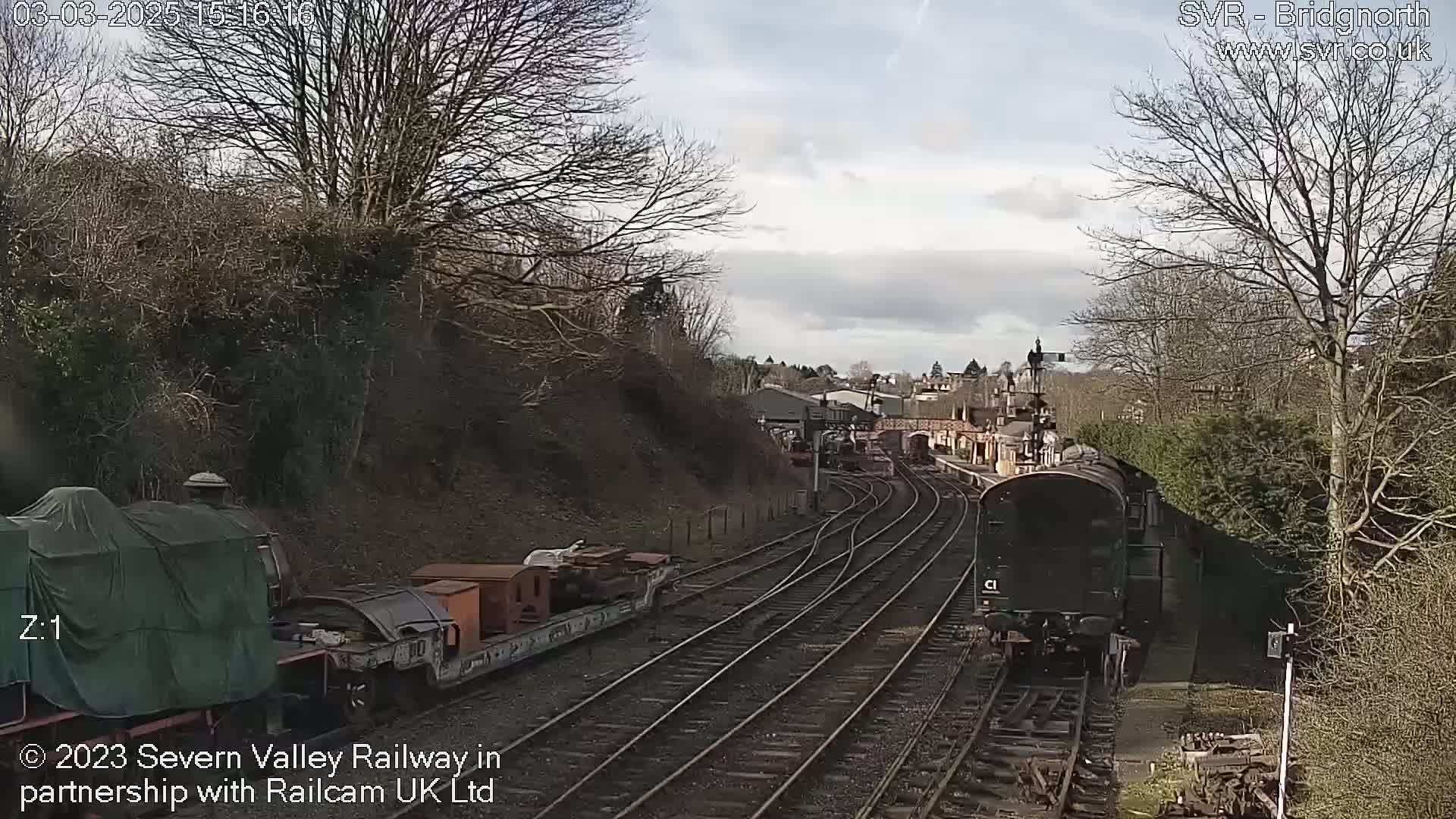 A train passes a railway station on a partly sunny day, with bare trees lining the tracks.