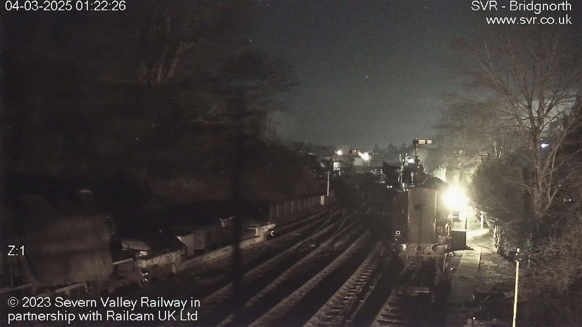 A nighttime, overhead view of a train station with train tracks and a partially visible train, under a clear, starry sky.